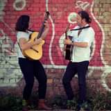 May be an image of 1 person, playing a musical instrument, standing, guitar, outdoors and brick wall