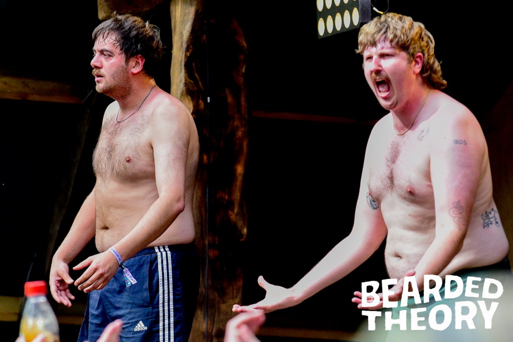 Two shirtless male performers energetically engaging with the audience on stage at a music festival.