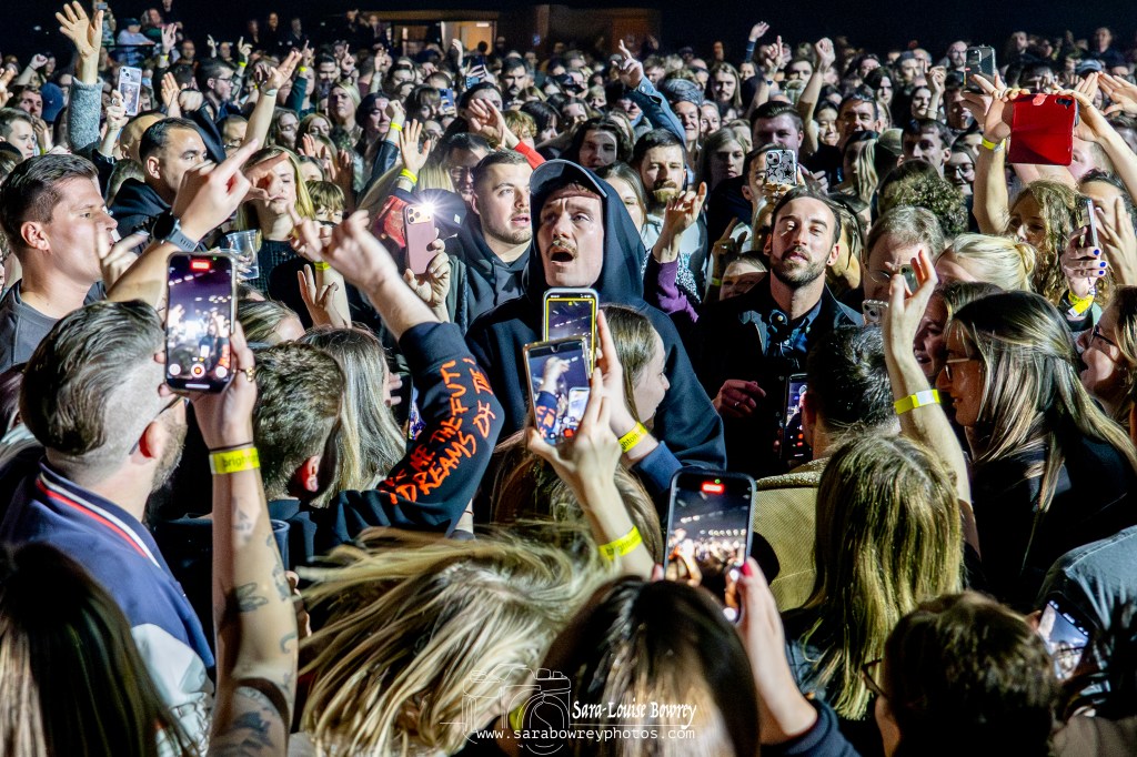 A crowded concert scene with enthusiastic fans holding up their phones, capturing the moment as an artist engages with the audience. The atmosphere is lively and electric.